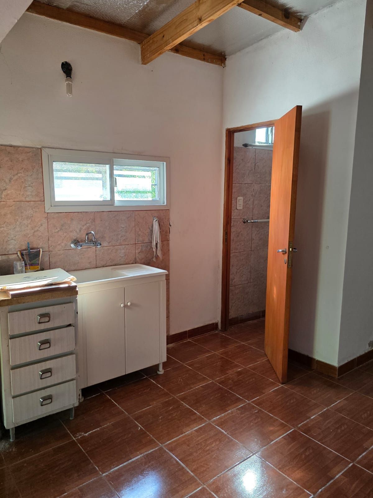 Pequeña cocina alicatada con fregadero, ventana, armarios blancos, vigas de madera en el techo y puerta abierta.