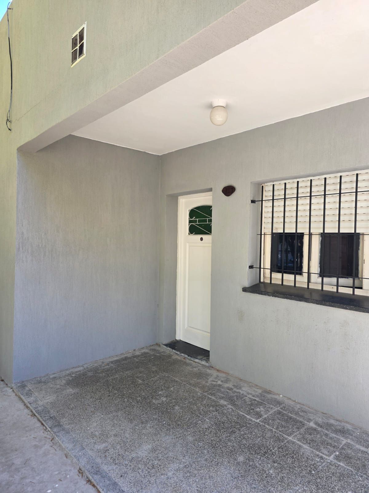 Entrada cubierta a un apartamento con paredes grises, puerta blanca y ventana con barrotes bajo un alero.