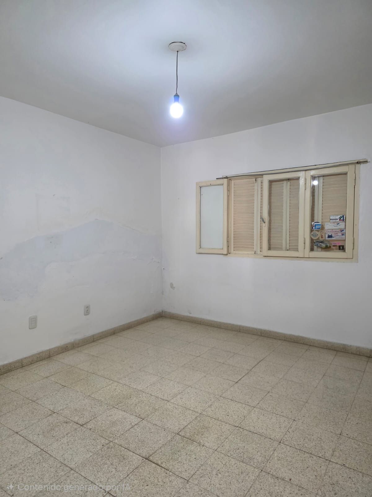 Habitación blanca vacía con suelo de baldosas, lámpara de techo y ventana con contraventanas.