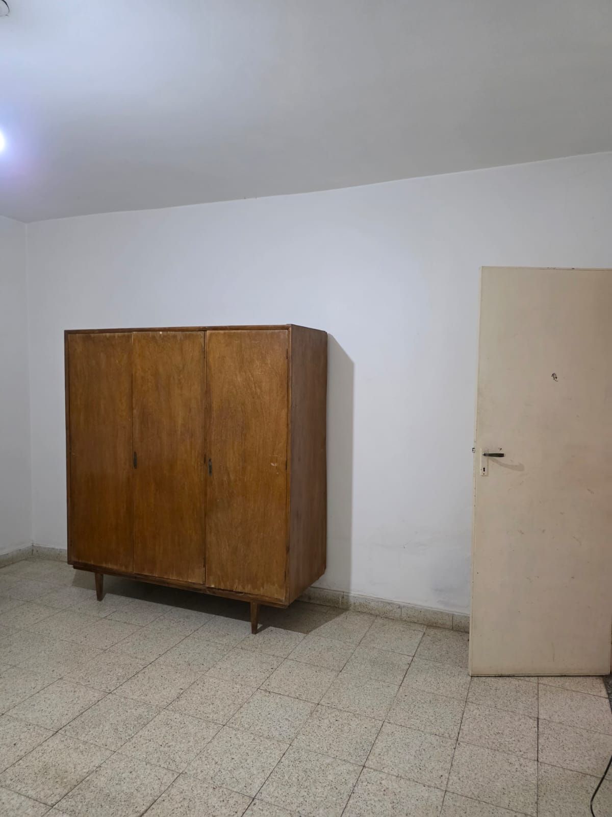 Habitación vacía con un gran armario de madera contra una pared blanca y una puerta beige cerrada.