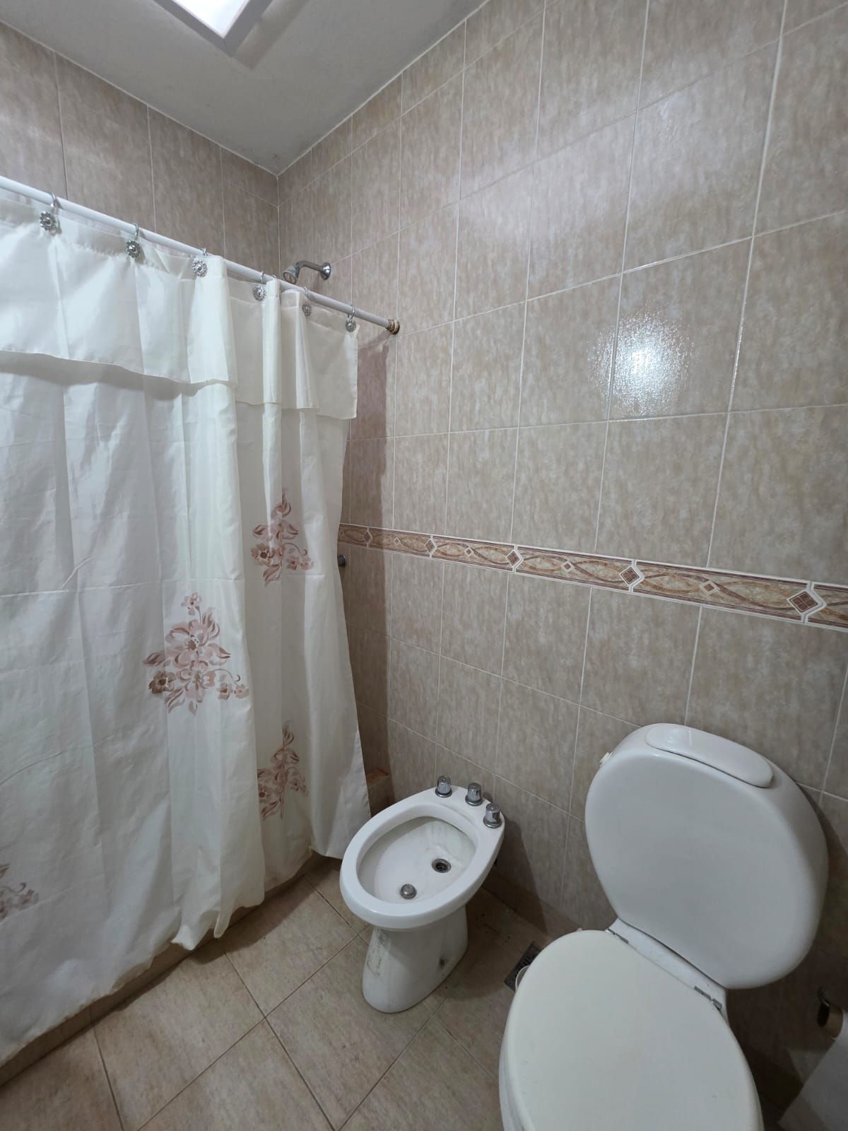 Baño pequeño con cortina de ducha, bidé e inodoro, con paredes revestidas de azulejos beige.