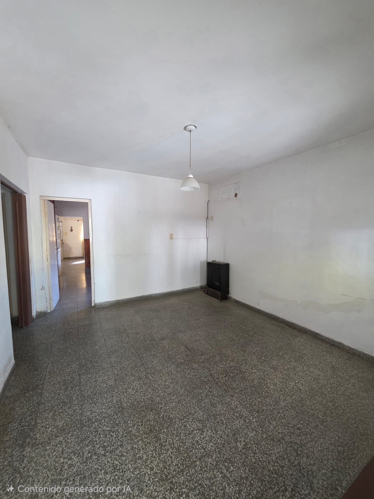 Habitación blanca vacía con suelo moteado, puerta que da al pasillo y una sola lámpara colgante.