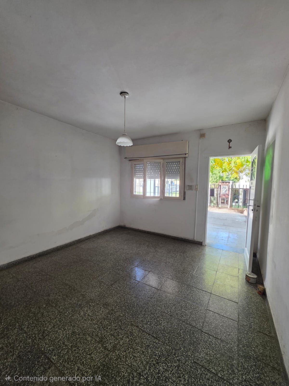 Habitación vacía con suelo de terrazo, paredes blancas, ventana, lámpara de techo y puerta abierta al exterior.