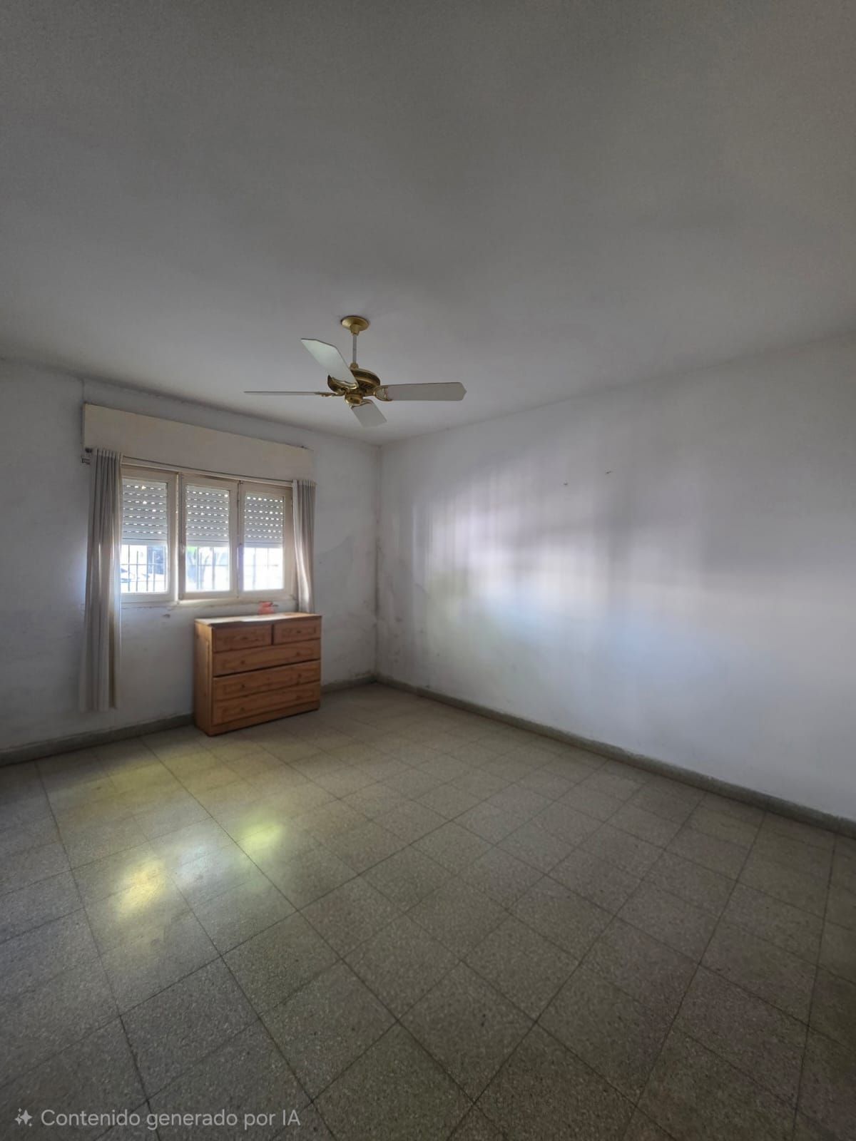 Habitación vacía con suelo de baldosas, paredes blancas, ventilador de techo y una cómoda de madera junto a la ventana.