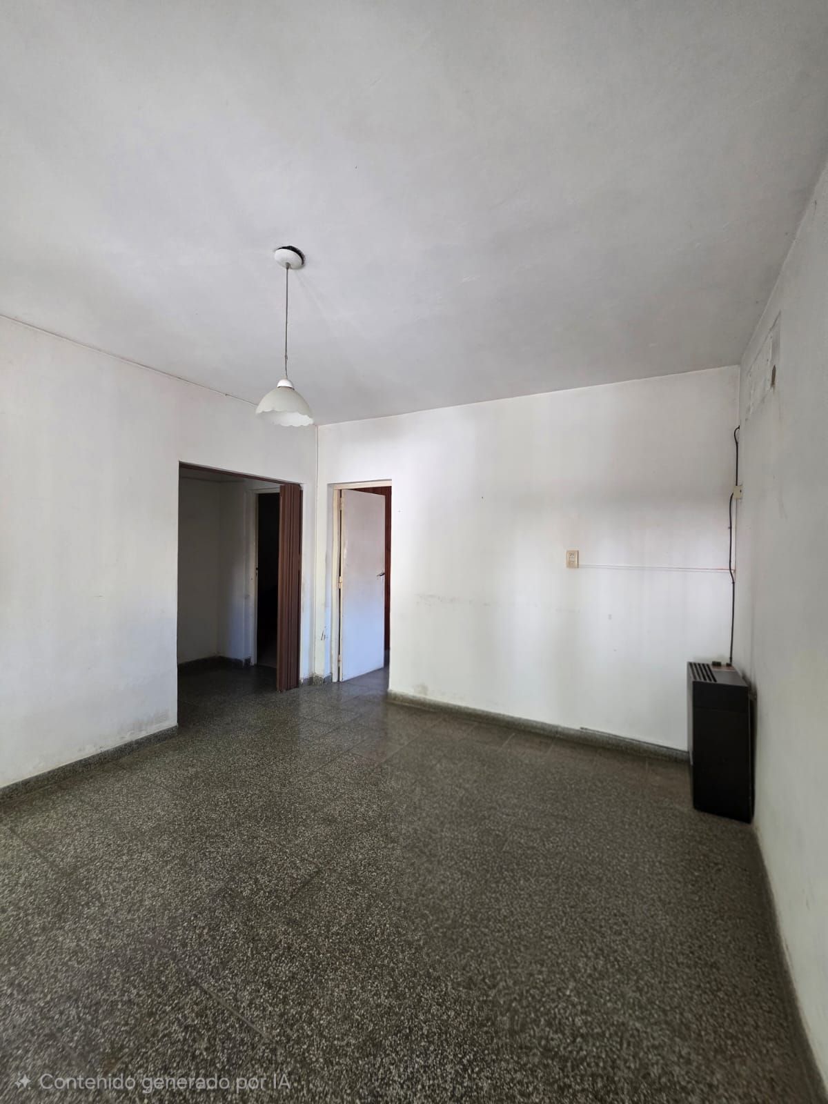 Habitación blanca vacía con suelo moteado, lámpara colgante y dos puertas abiertas.