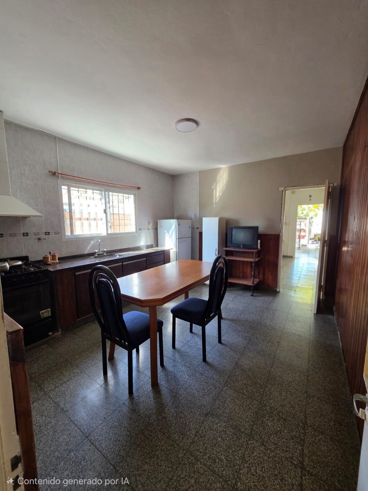 Cocina-comedor vacía con armarios oscuros, mesa y sillas, nevera y un pequeño televisor en la esquina.