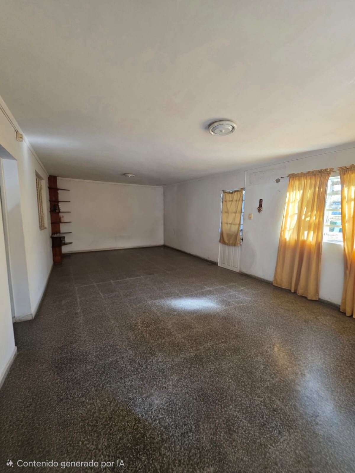 Sala de estar vacía con alfombra marrón, paredes blancas y luz solar que se filtra a través de cortinas amarillas.