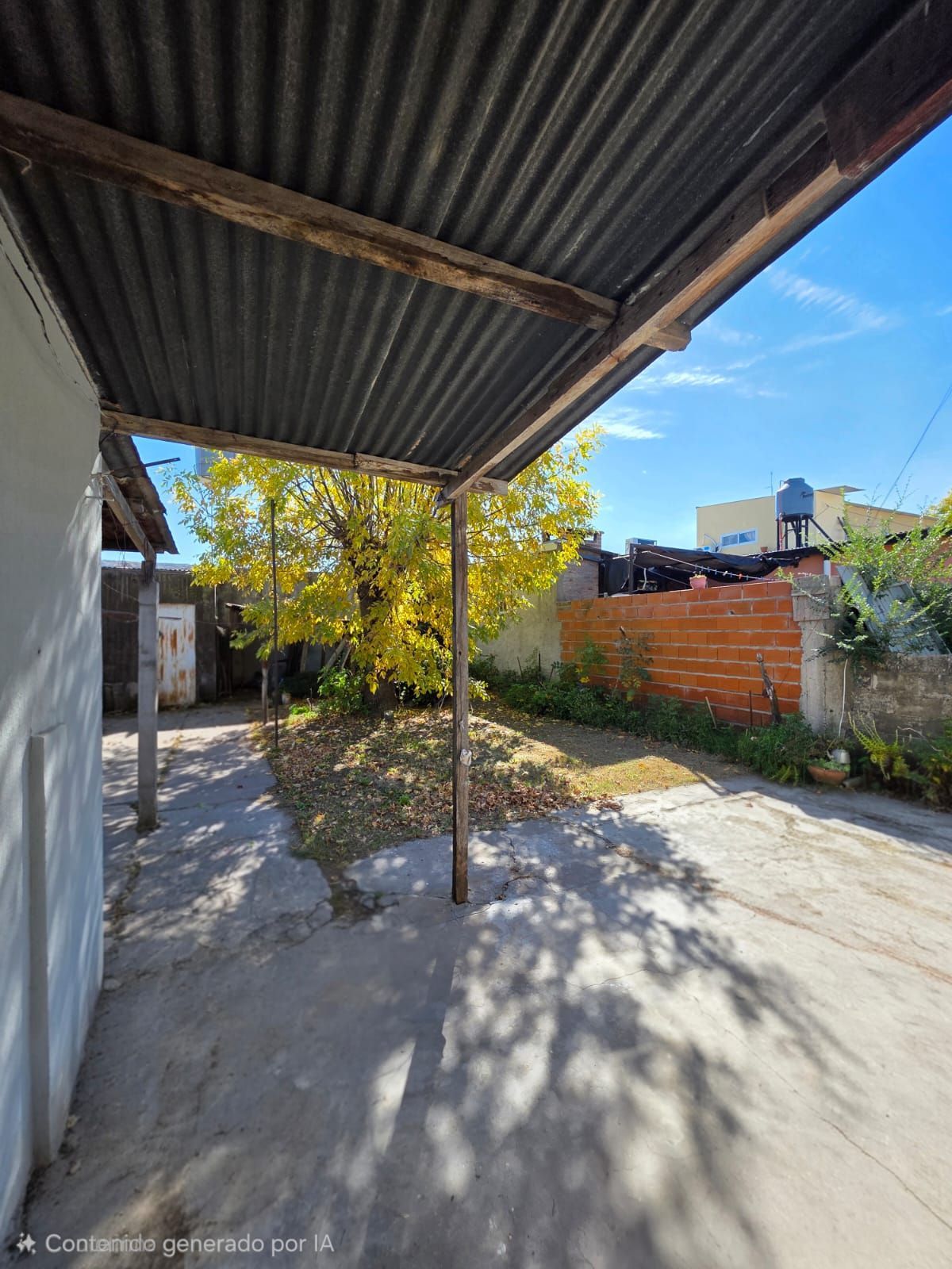 Patio cubierto de hormigón con techo ondulado, árbol central y muro de ladrillo junto a un jardín soleado.