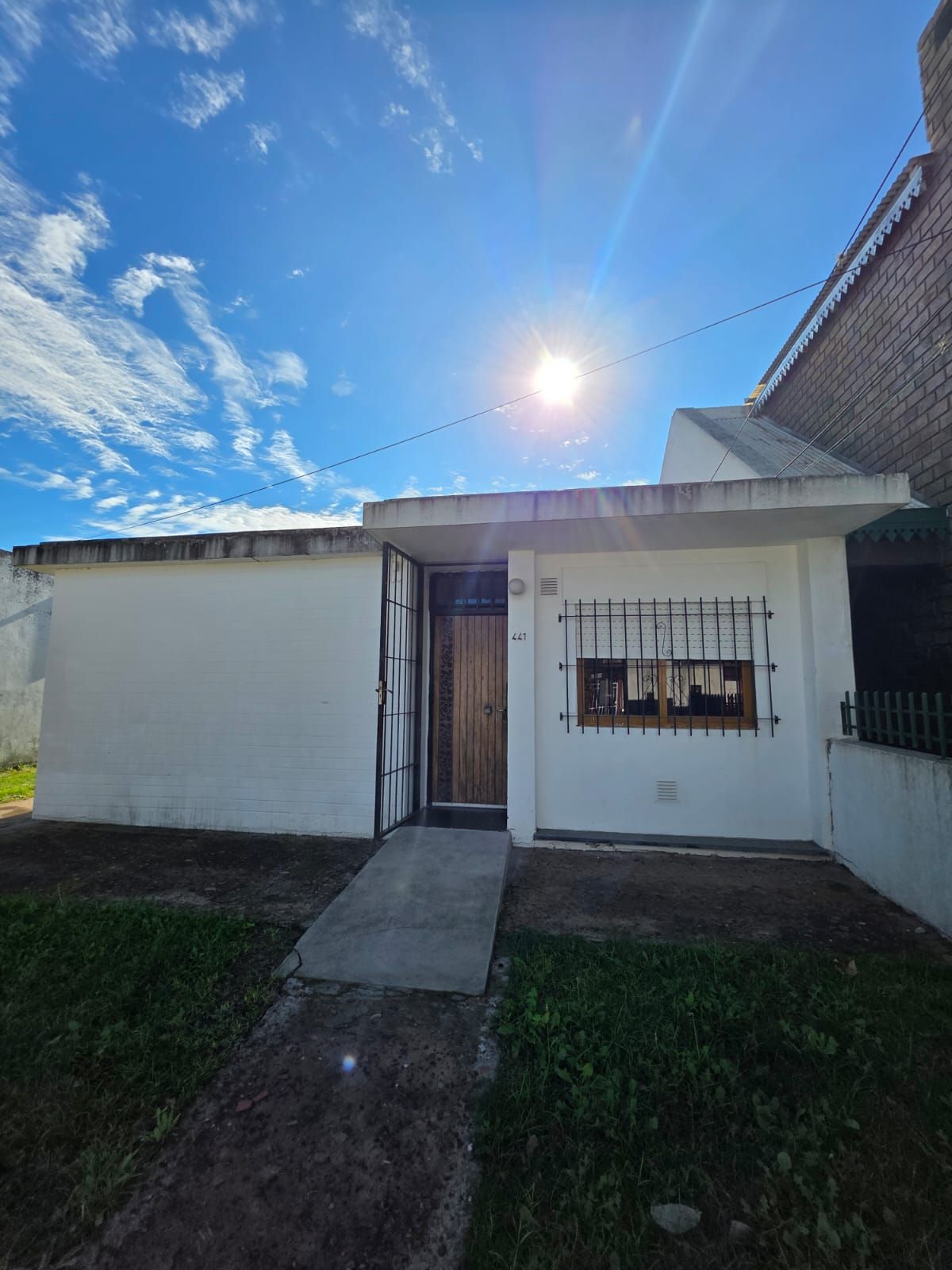 Pequeña casa blanca con un camino de entrada, una ventana enrejada y un sol brillante en un cielo azul.