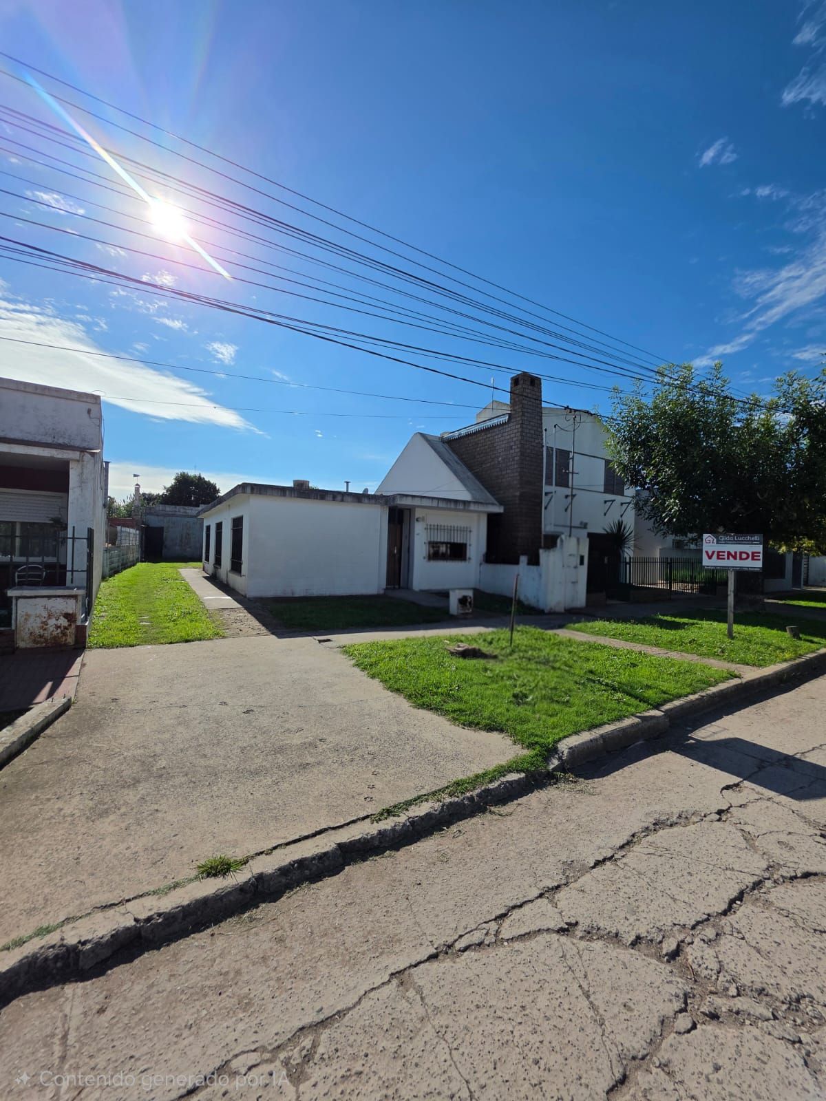 Calle residencial soleada con una casa blanca, entrada para vehículos, césped verde y líneas de servicios públicos aéreas.