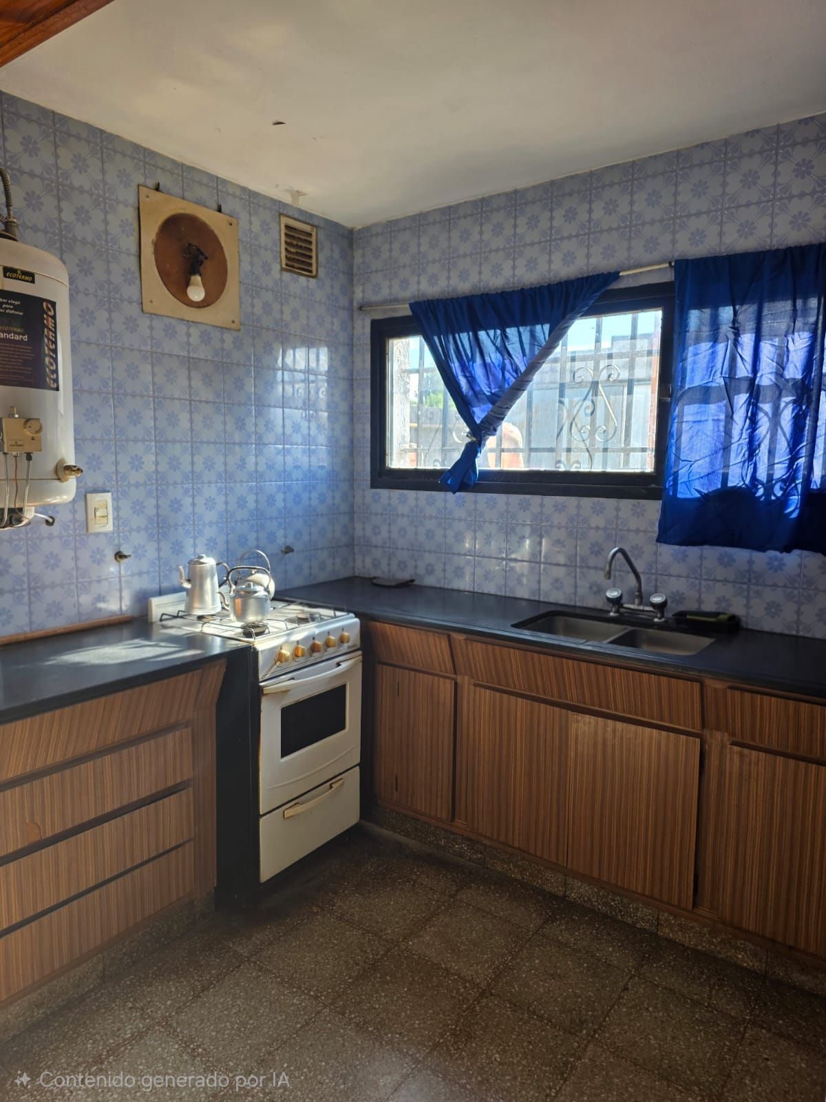 Cocina compacta con paredes estampadas en azul, armarios de madera, estufa, fregadero y ventana con cortinas azules.