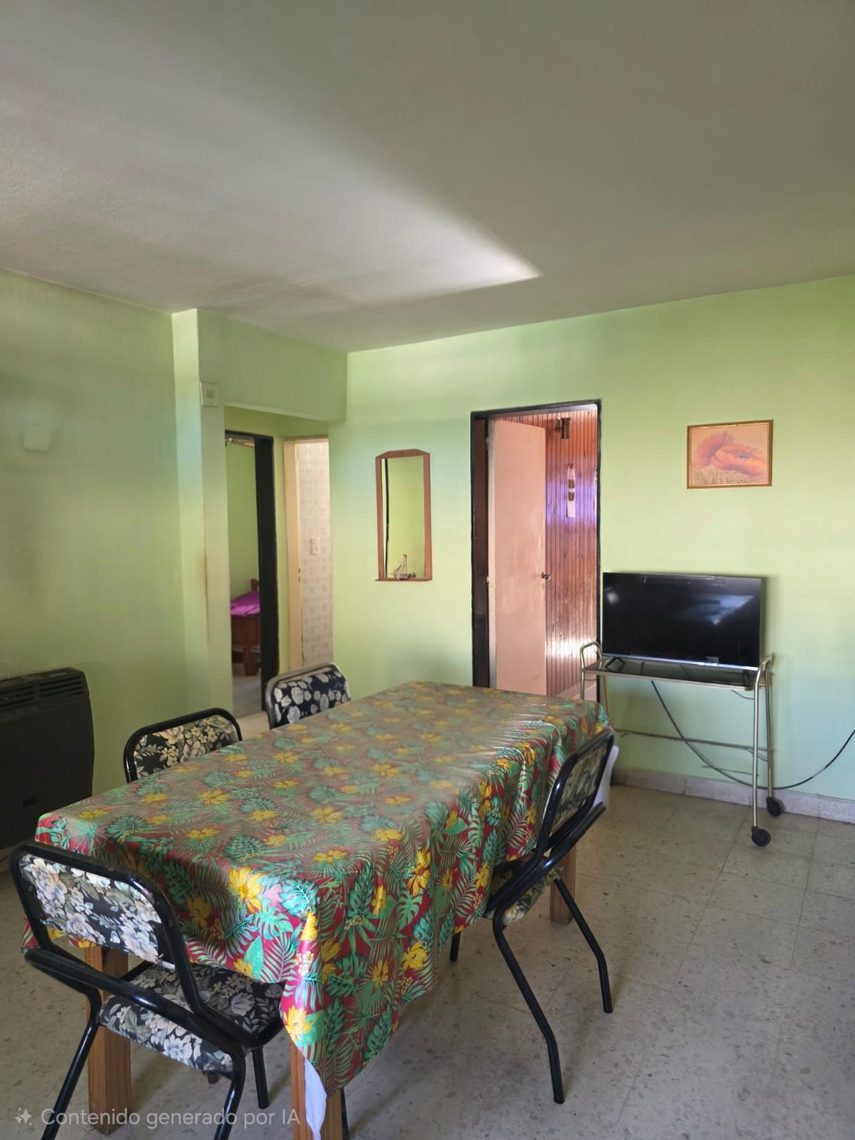 Comedor con mantel estampado, sillas, televisor y puerta abierta en una habitación de color verde pálido.