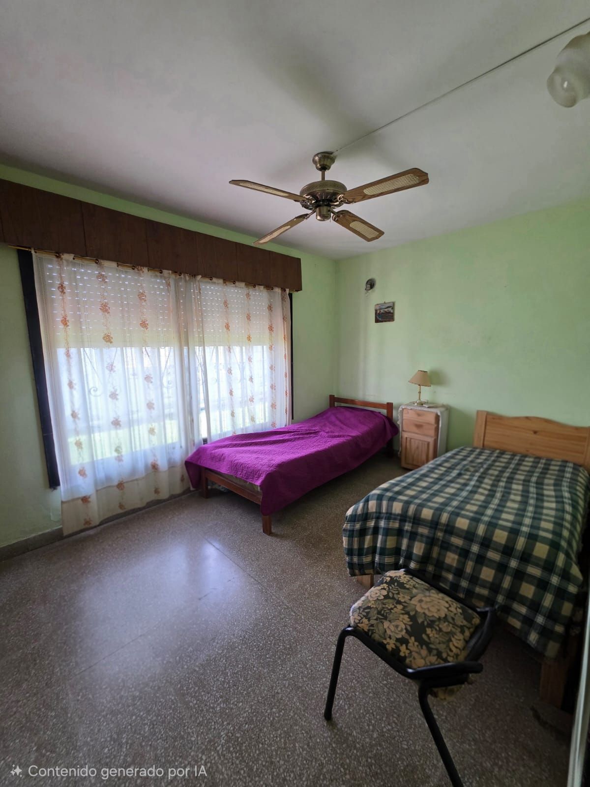 Habitación pequeña con dos camas individuales, paredes verdes y un ventilador de techo cerca de una ventana con cortinas.