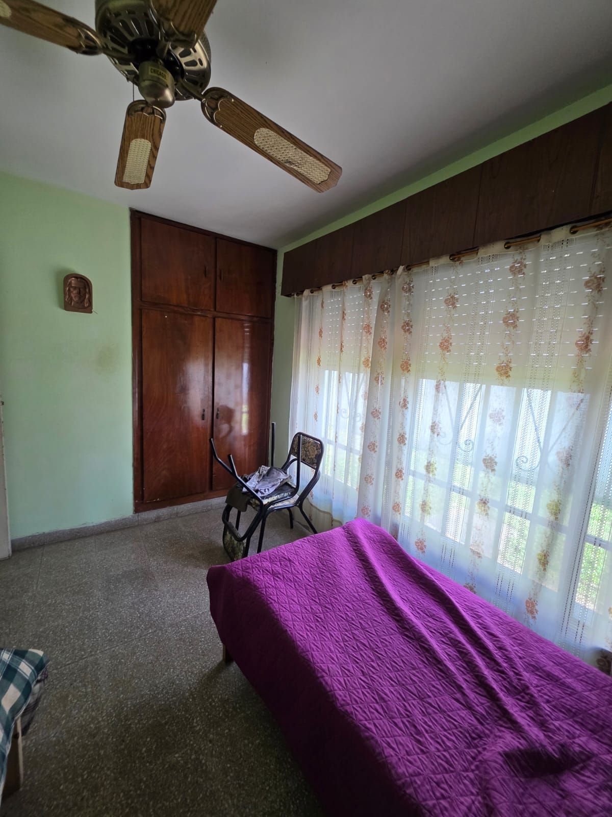 Habitación pequeña con colcha morada, ventana grande con cortinas, armario, silla y ventilador de techo.