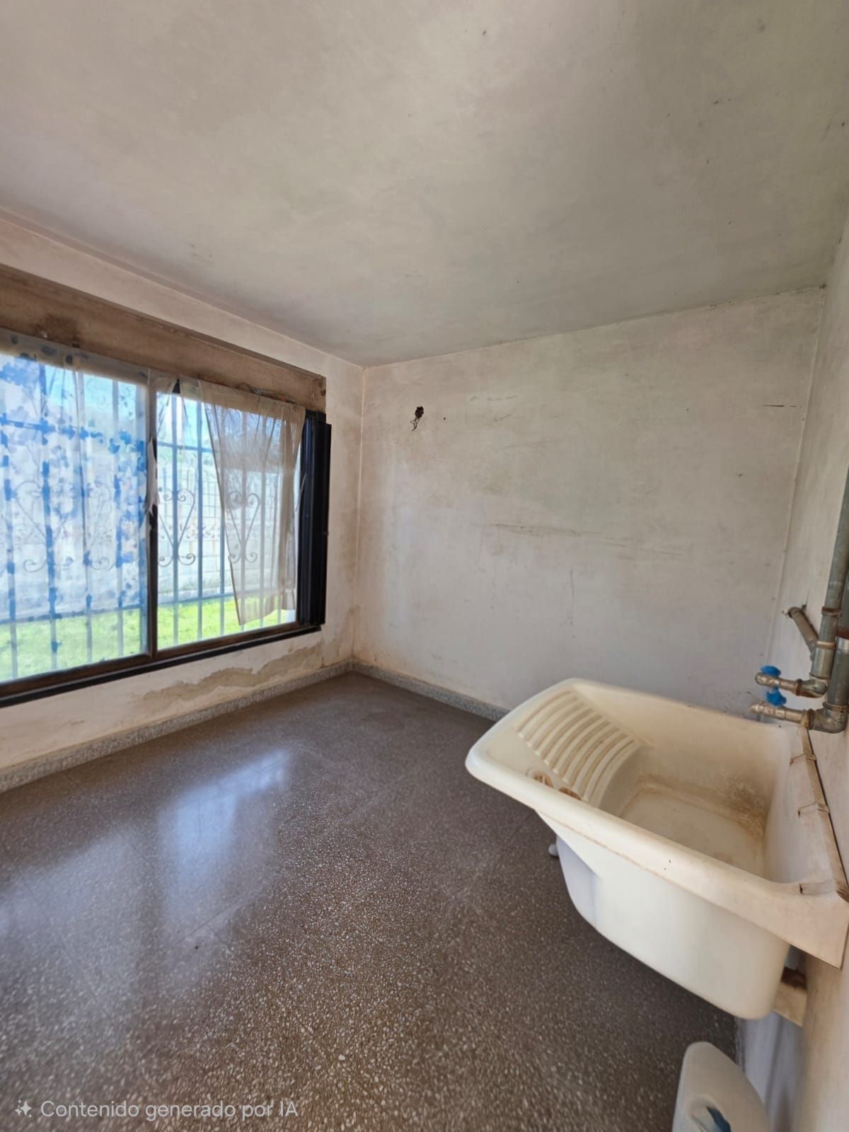 Habitación pequeña y vacía con una ventana enrejada y un fregadero blanco contra una pared lisa.