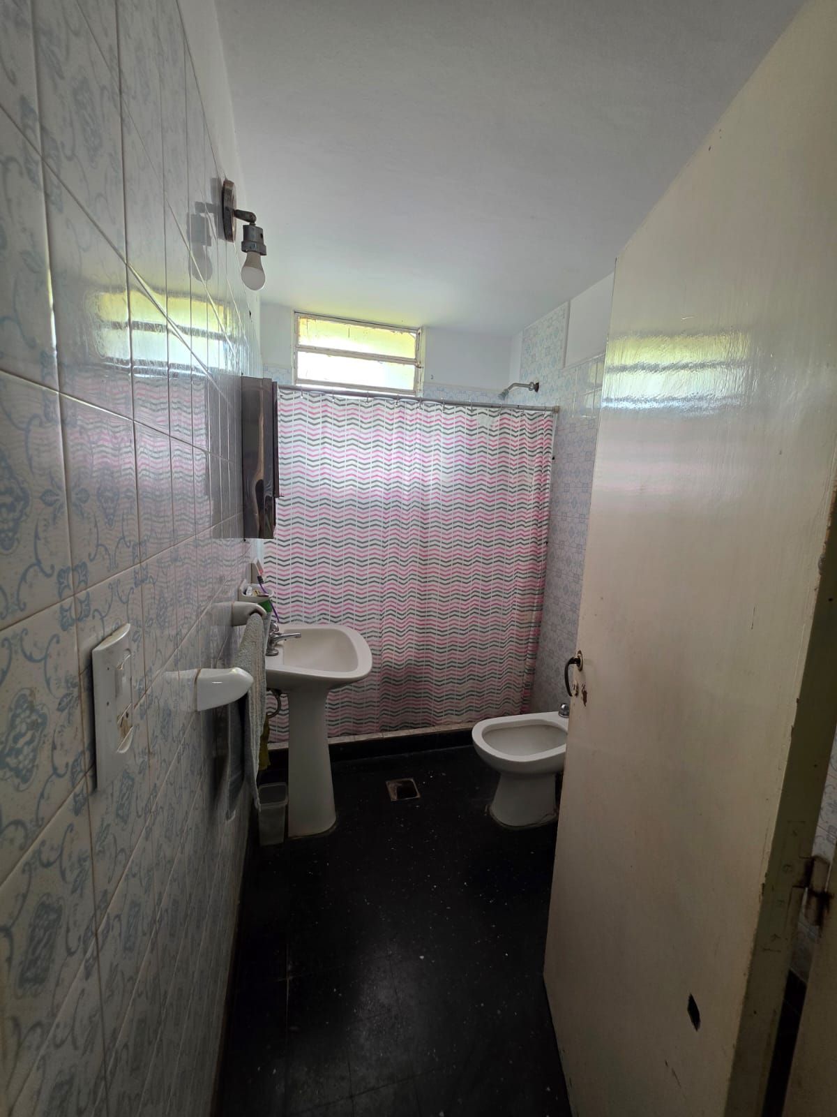 Pequeño baño público con azulejos, dos inodoros, un lavabo y suelo negro.