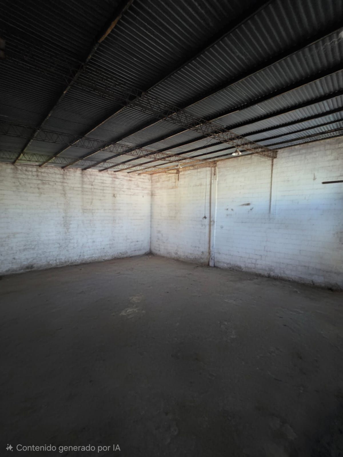 Habitación industrial vacía con paredes de ladrillo blanco, techo de chapa ondulada oscura y suelo de hormigón.