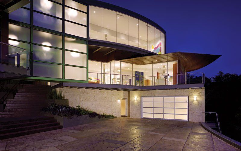 Modern curved glass home with lit interior at dusk; garage below.