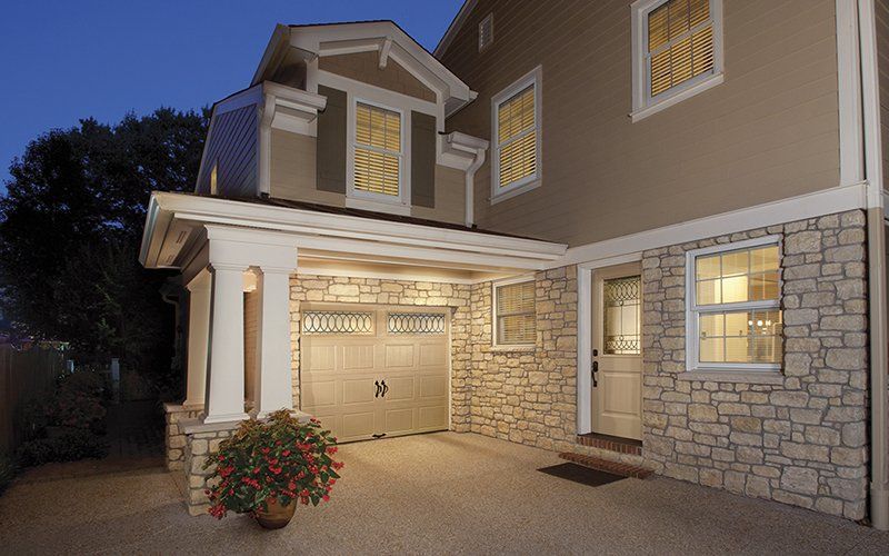 Beige two-story house with stone accents, garage, porch, and lit windows at dusk.