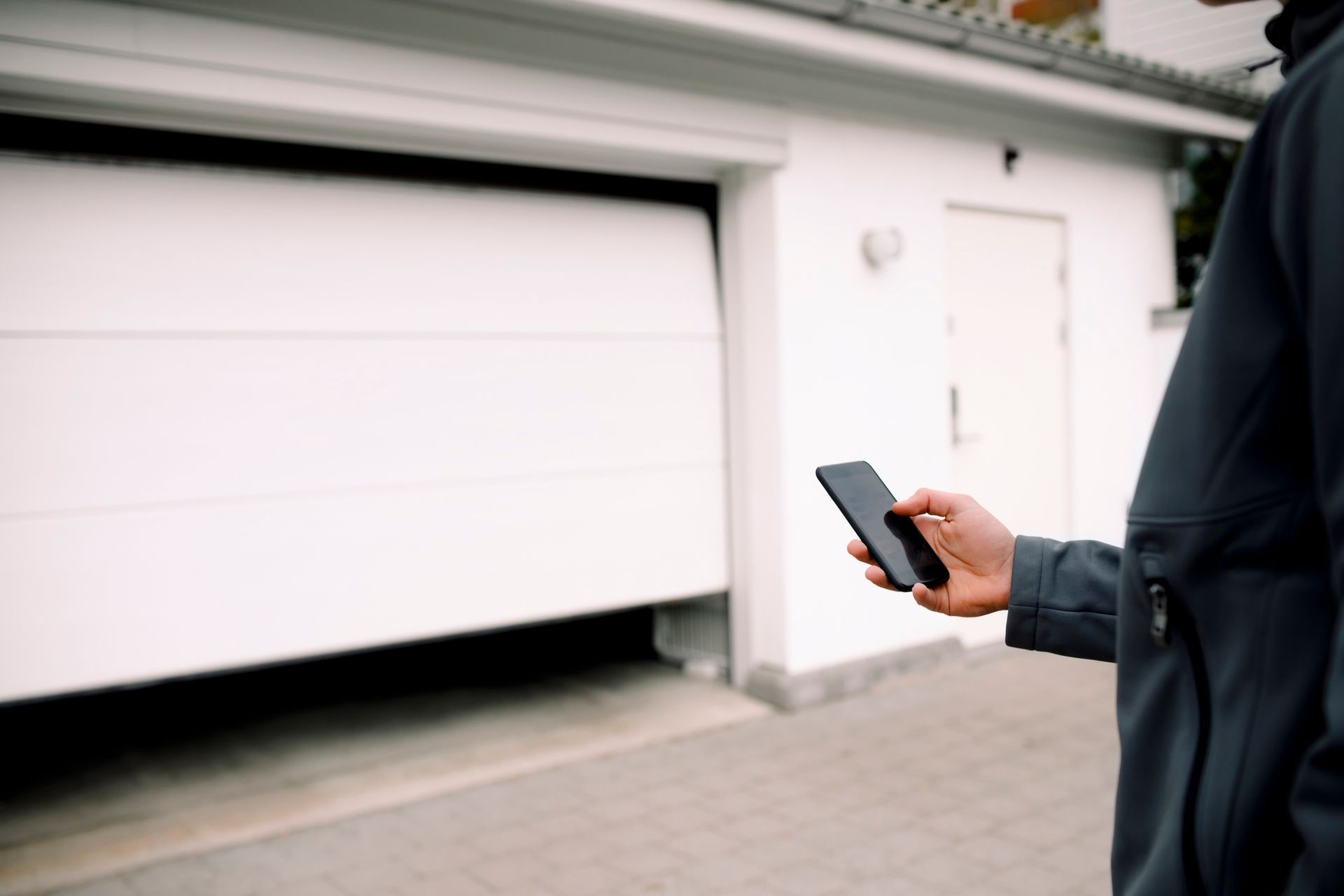 Person using a smartphone to open a white garage door. Exterior shot, brick driveway.