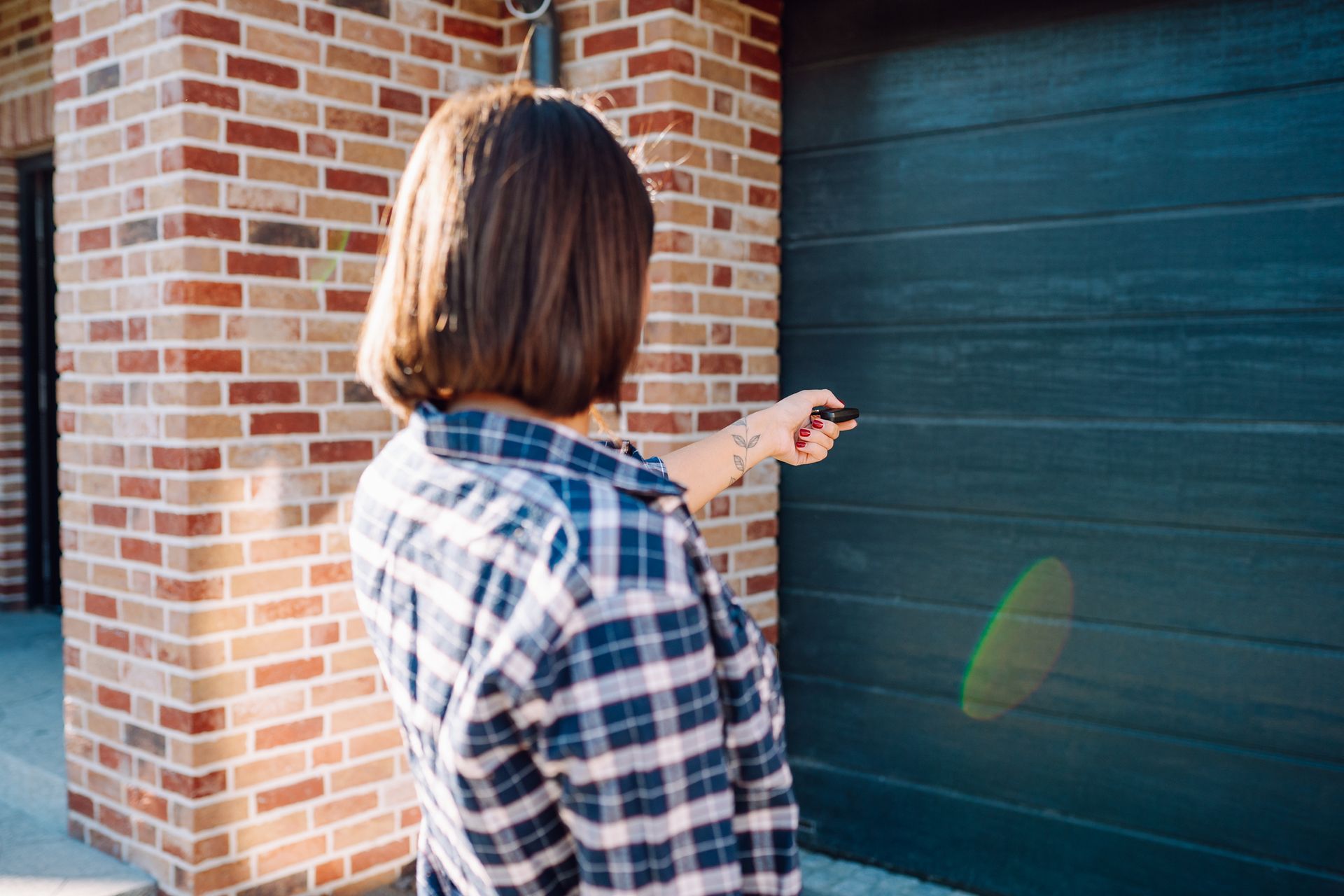 Woman using a remote to open a garage door. She stands in front of a blue-gray door next to a brick wall.