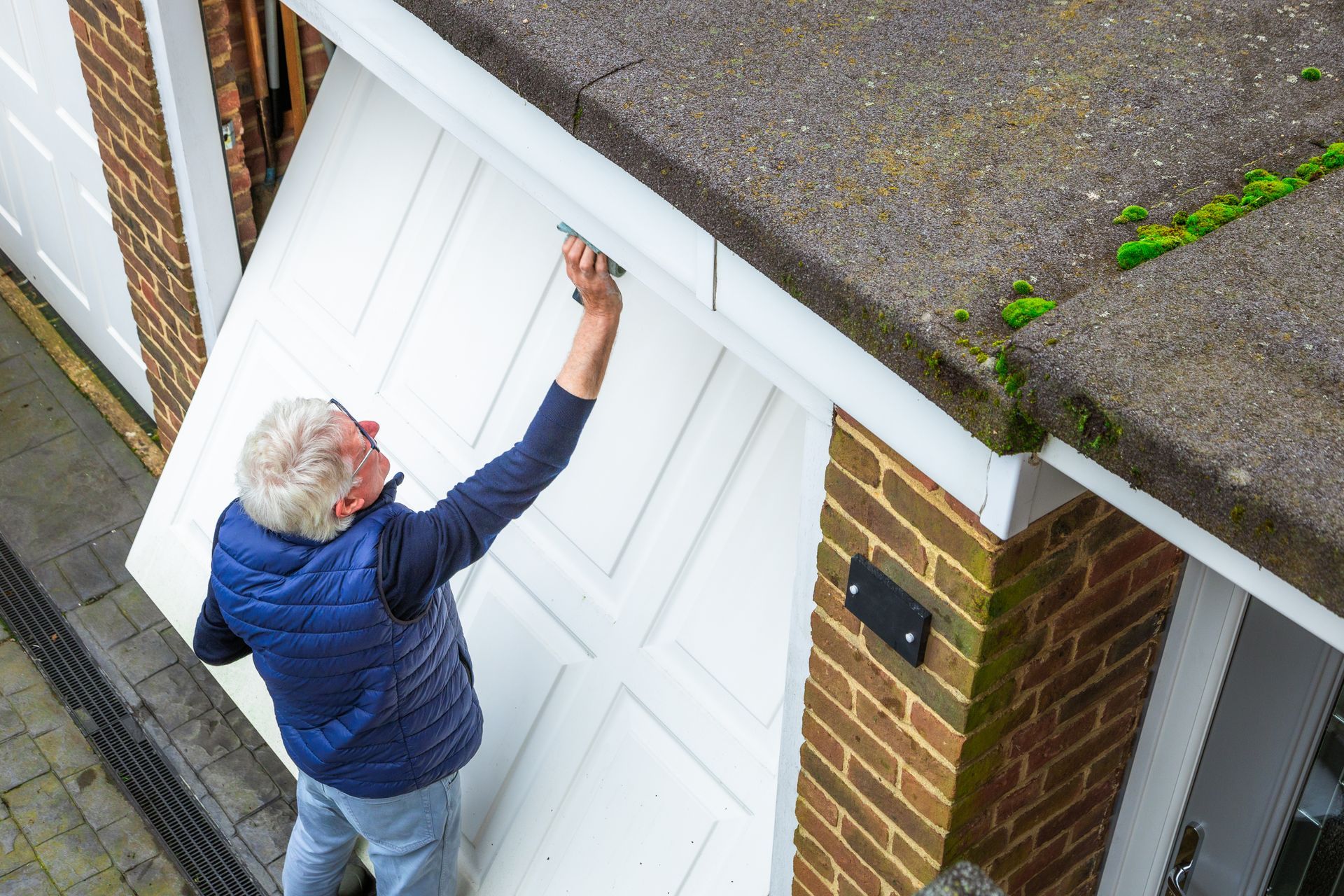 Man in blue vest opening a white garage door, next to a brick building.