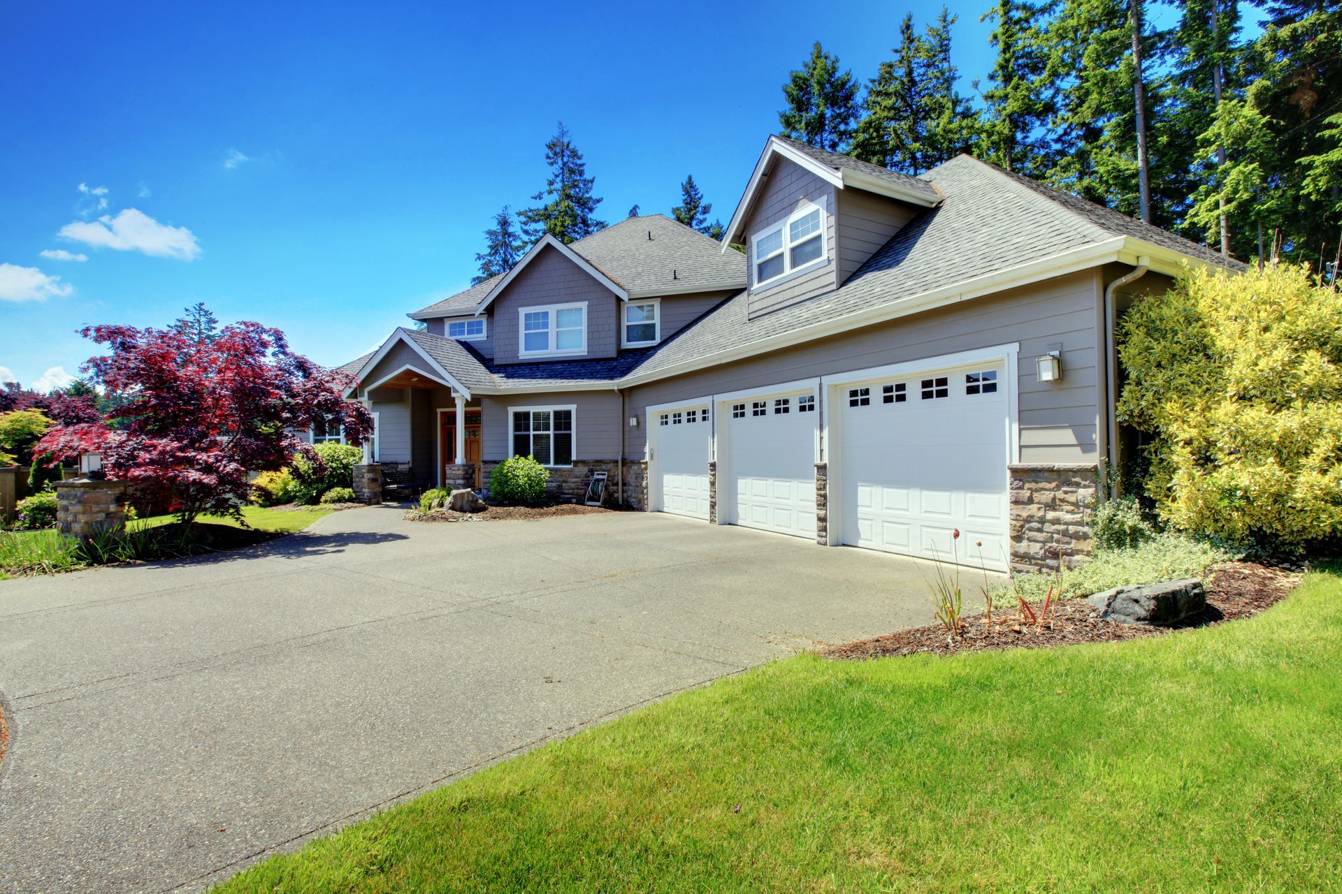 Gray house with three-car garage, driveway, and lush green lawn under a bright blue sky.
