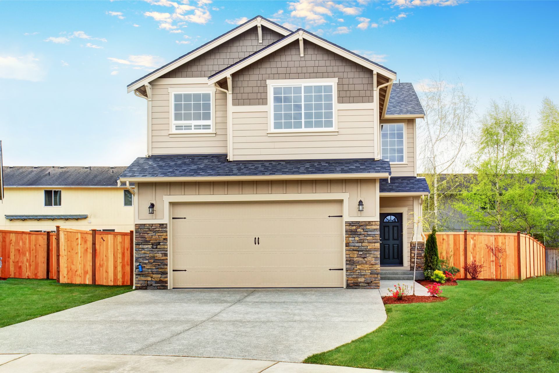 Two-story beige house with a tan garage door, set on a green lawn with a wooden fence.