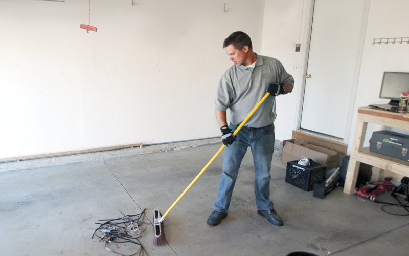 Man sweeping a garage floor with a broom.  He is wearing a grey shirt, jeans, and gloves.  The garage has a concrete floor.