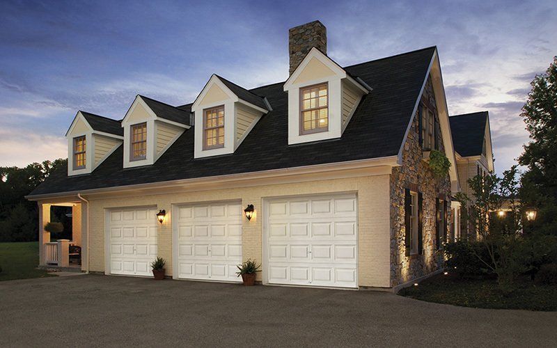 Three-car garage with white doors, tan facade, and stone accent. Three dormer windows. Evening light.