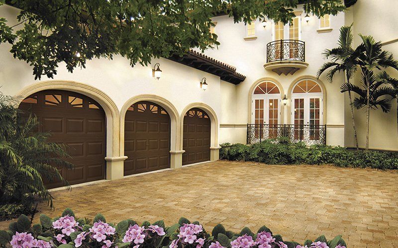 Brown garage doors under arched openings of a beige stucco house with a brick driveway and flowering bushes.