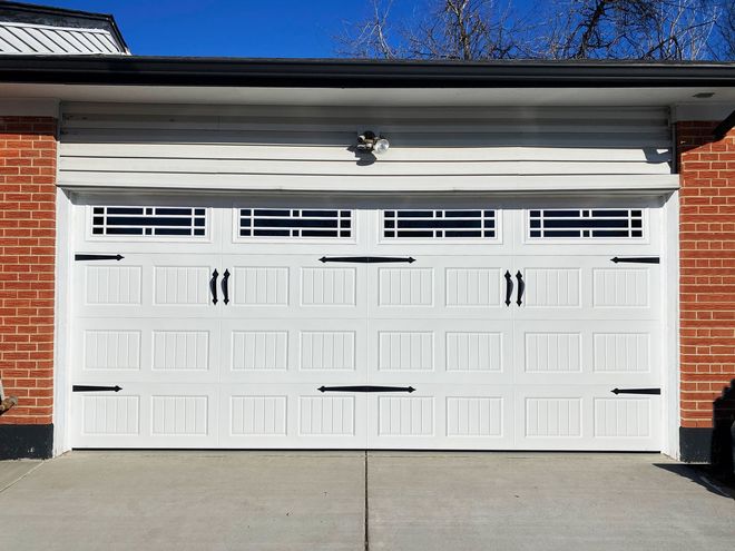 White garage door with decorative black hardware and windows, set in a brick and siding facade.