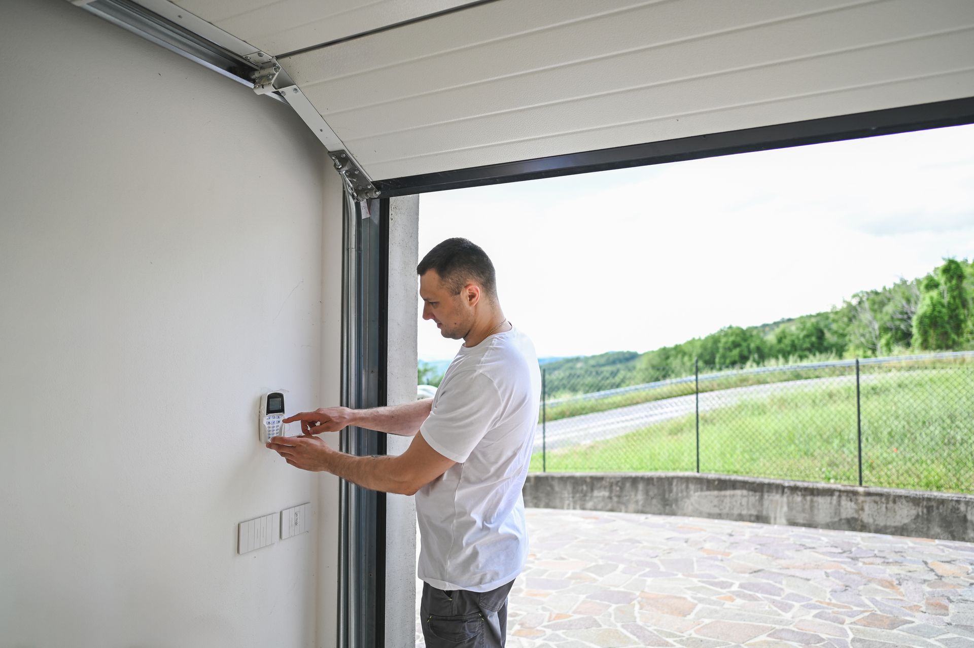 Man operates garage door opener on white wall, with open door revealing outdoor view.