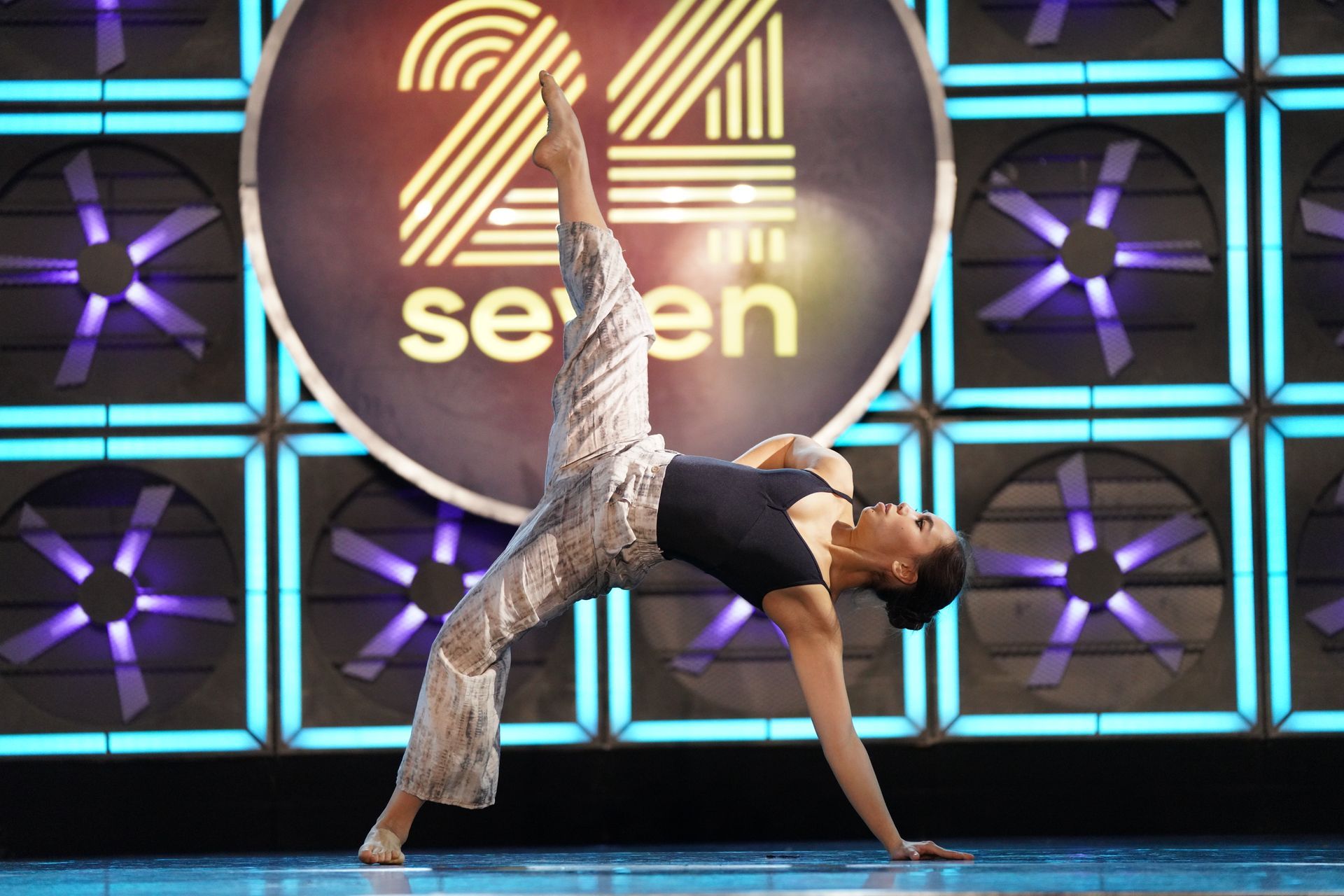 A dancer in a black top and patterned pants performs a high-leg extension on stage in front of a