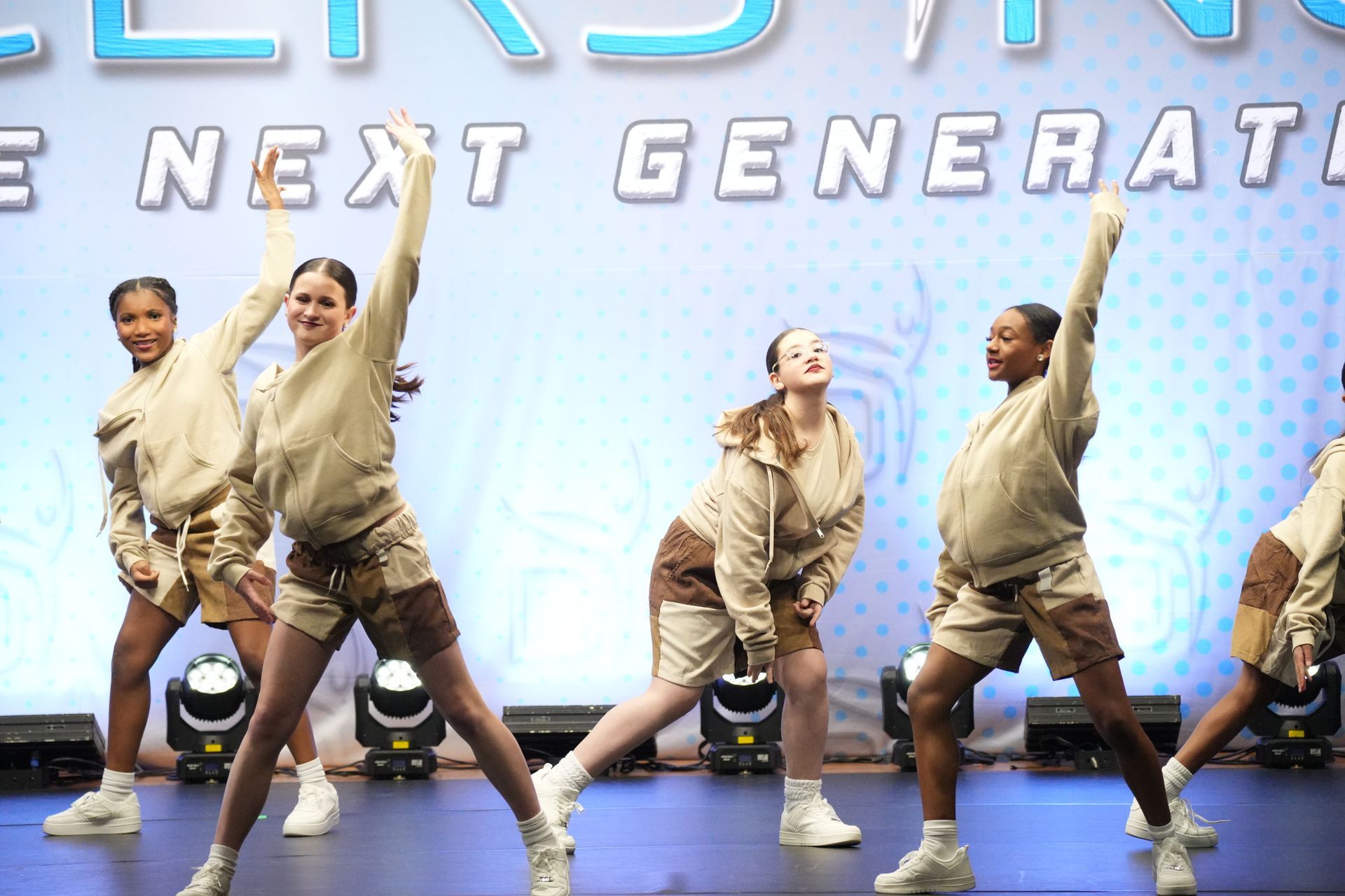 Dancers in matching beige hoodies and shorts perform on a stage with a light blue backdrop. E-Street Dance Academy, Edgewood, MD