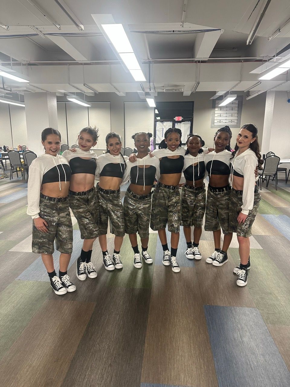 Eight dancers in matching white cropped hoodies and camouflage shorts stand in a line in a room with overhead lighting. E-Street Dance Academy, Edgewood, MD