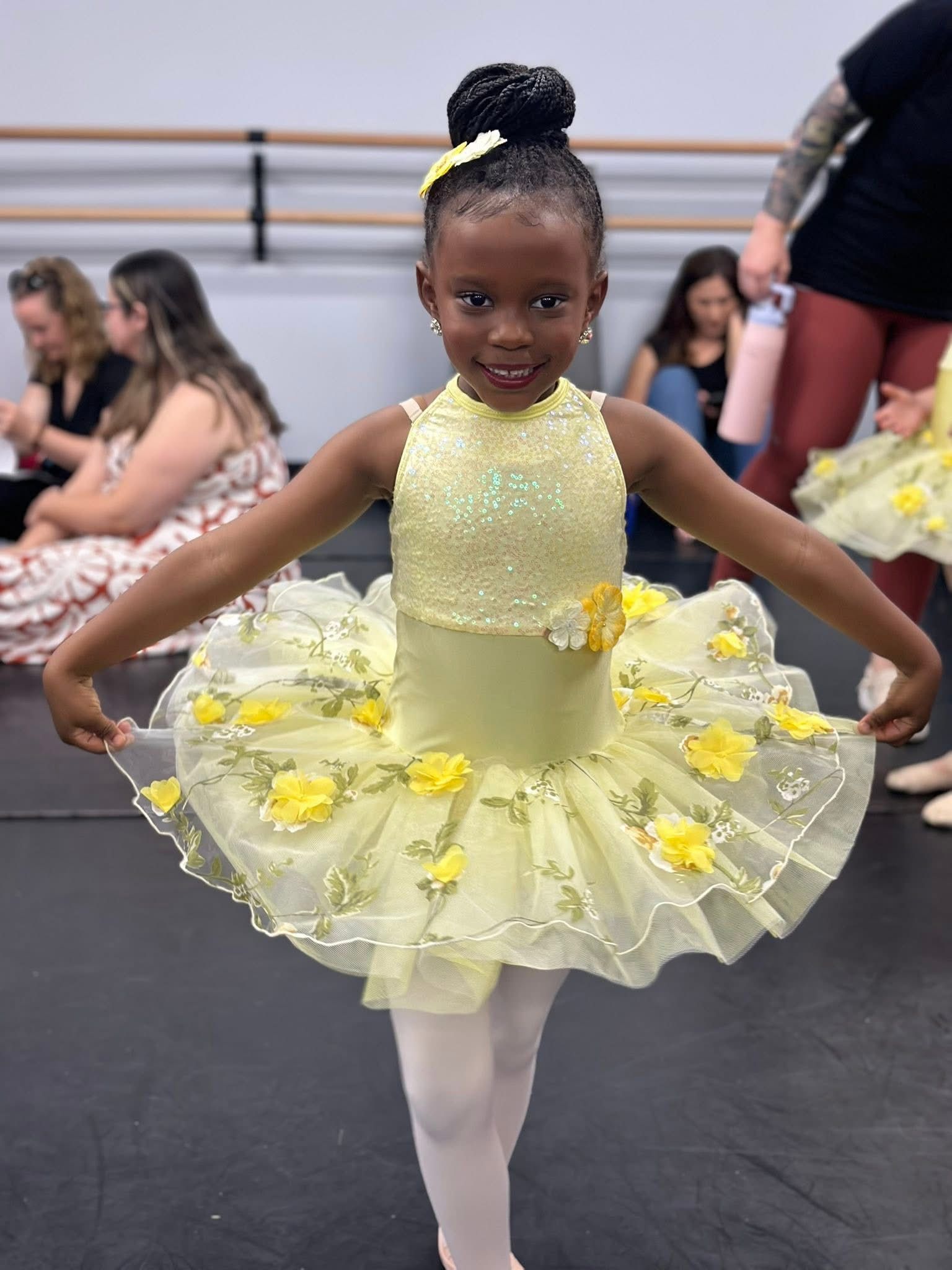A smiling child in a yellow ballet tutu with floral details poses in a dance studio with ballet barres in the background. E-Street Dance Academy, Edgewood, MD