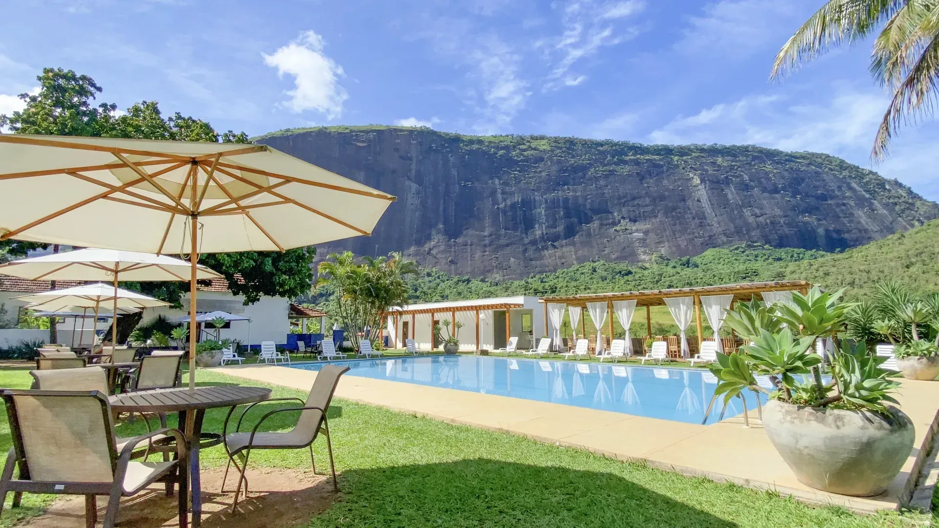 Uma piscina com uma montanha ao fundo e mesas e cadeiras sob guarda-sóis