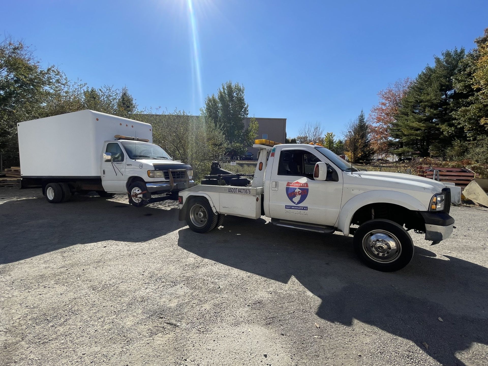White tow truck towing a box truck on gravel under a blue sky.