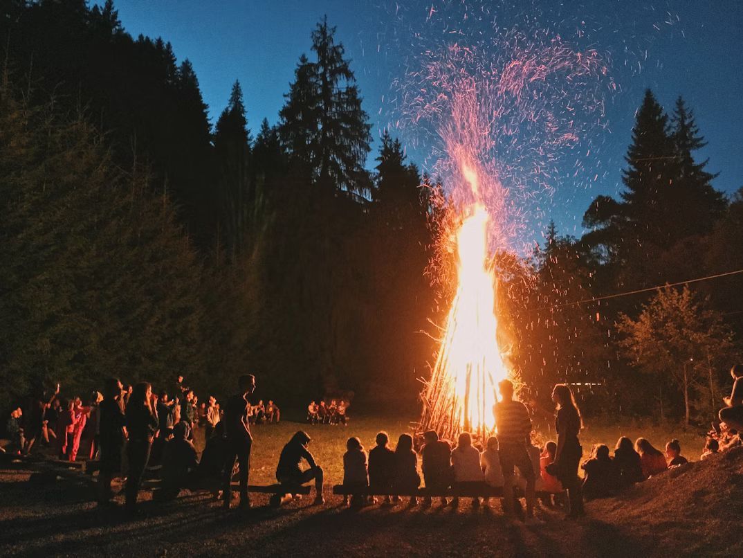 Large bonfire with sparks, people gathered around sitting on benches at night in a forest.