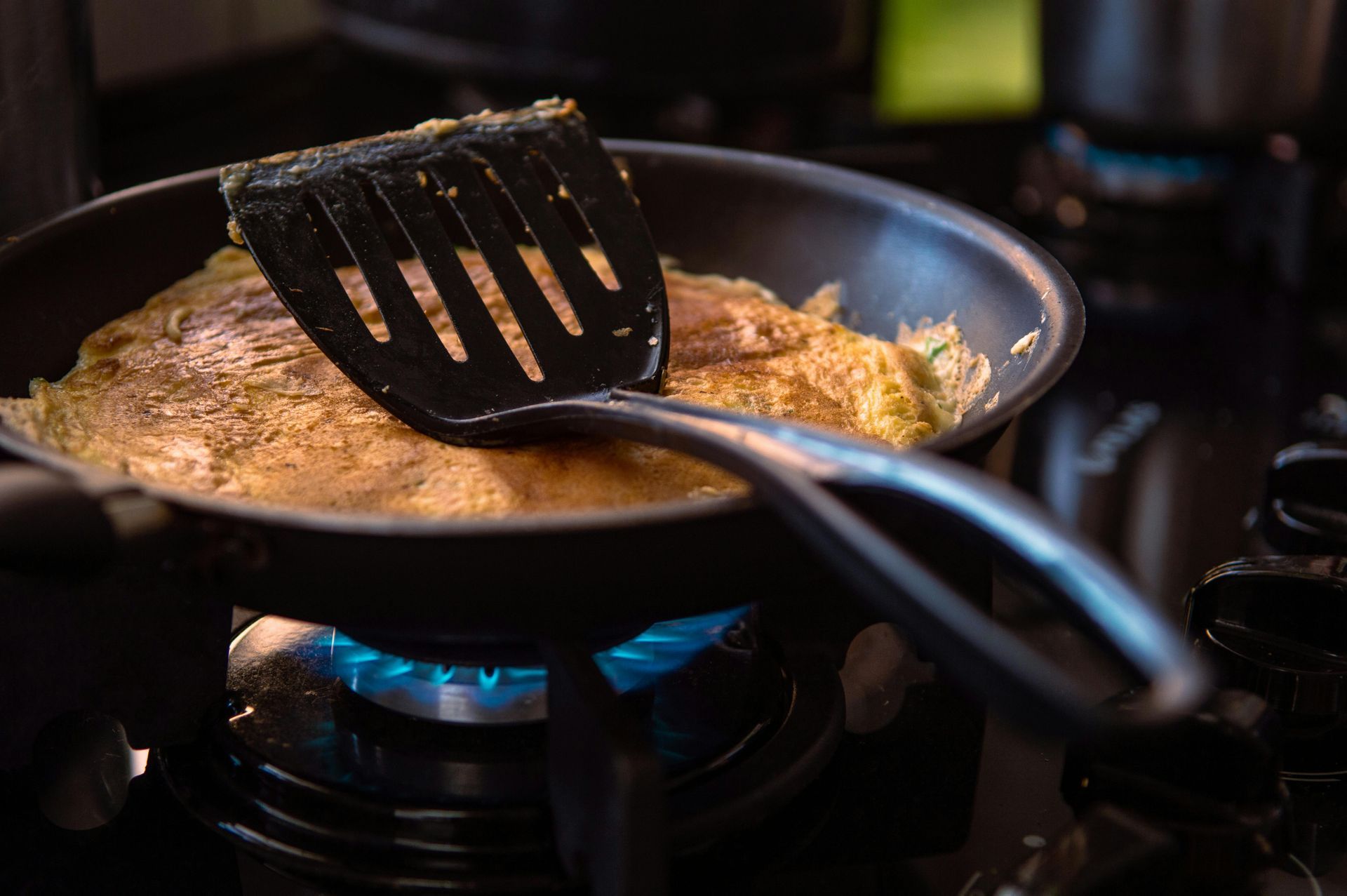 An omelet cooking in a pan on a gas stove, being flipped with a black spatula.