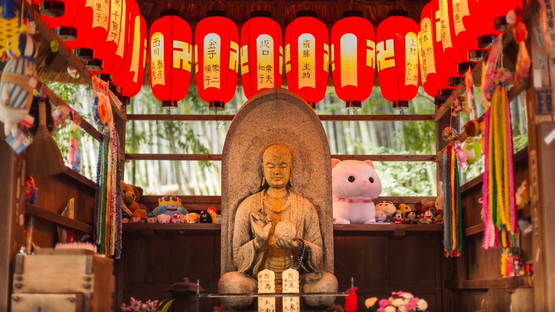 Stone Buddha statue in a shrine, red lanterns overhead, with colorful decorations and a plush toy.
