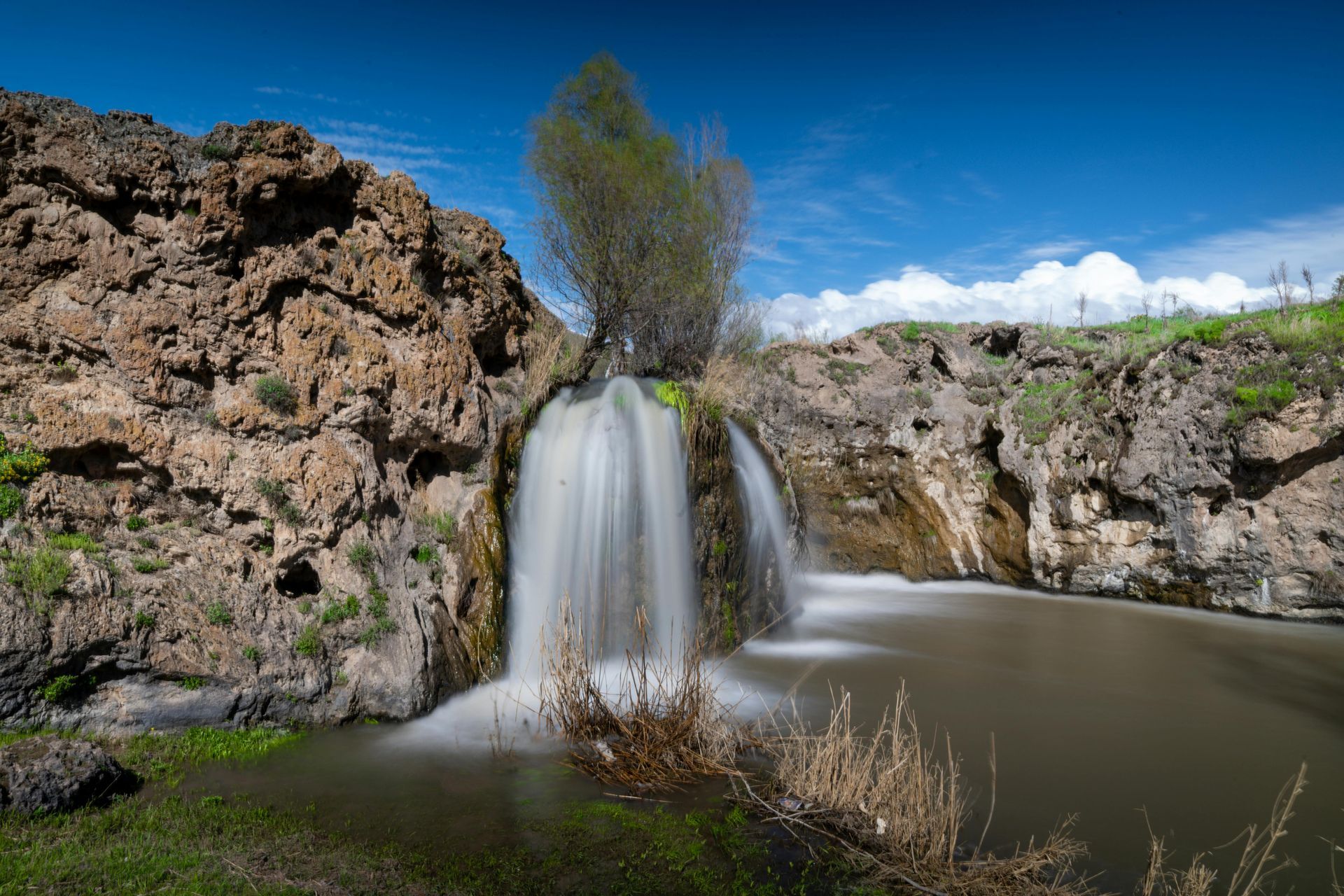 A waterfall is surrounded by rocks and grass in the middle of a river.