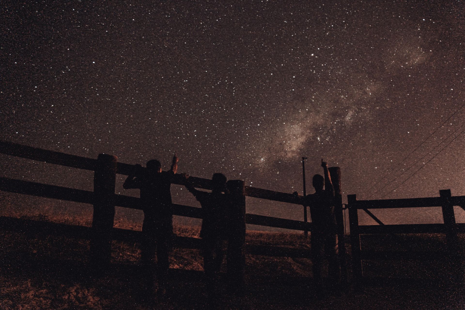 Silhouettes of people looking up at a starry night sky, near a wooden fence.