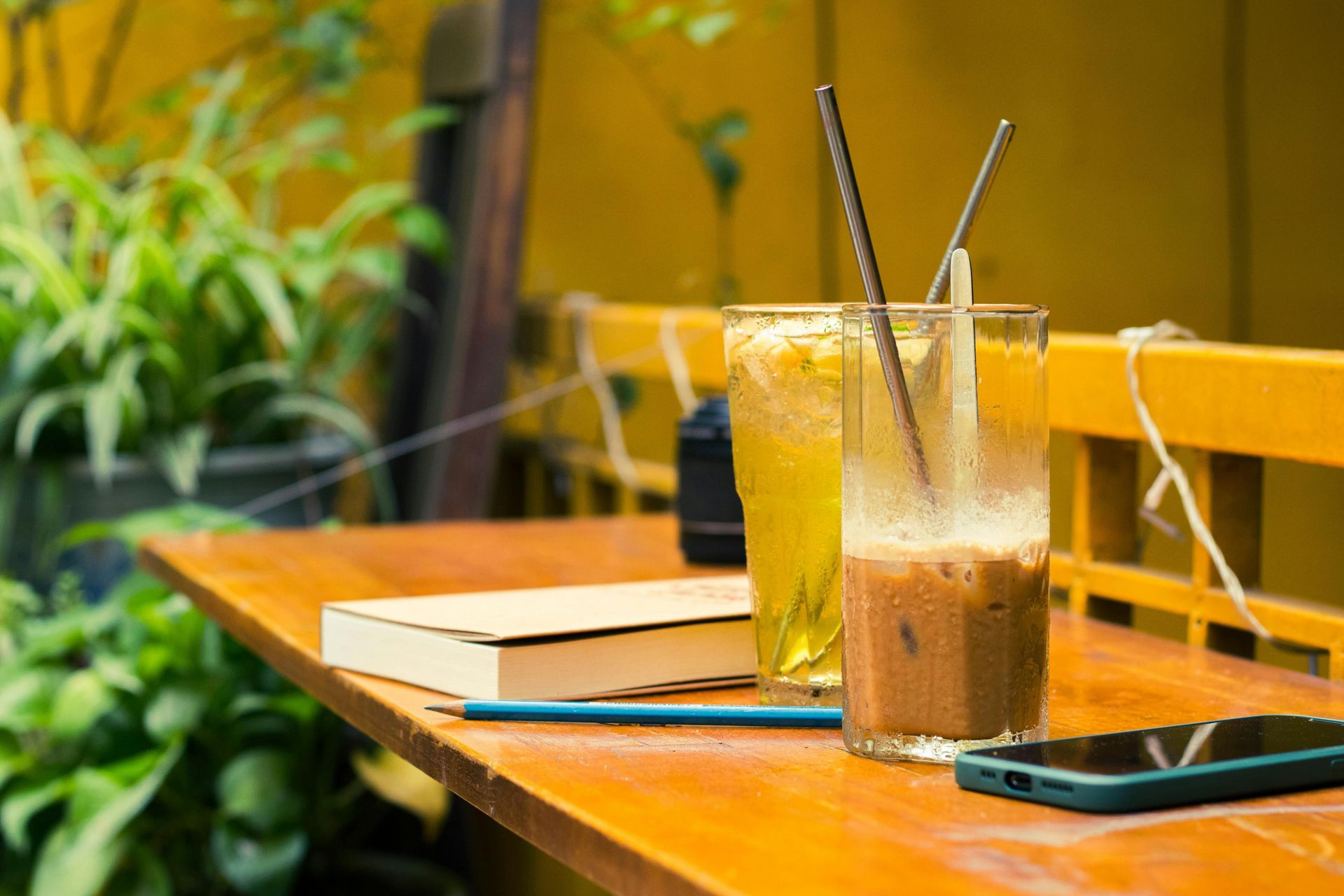 A table with two drinks , a book , and a cell phone on it.