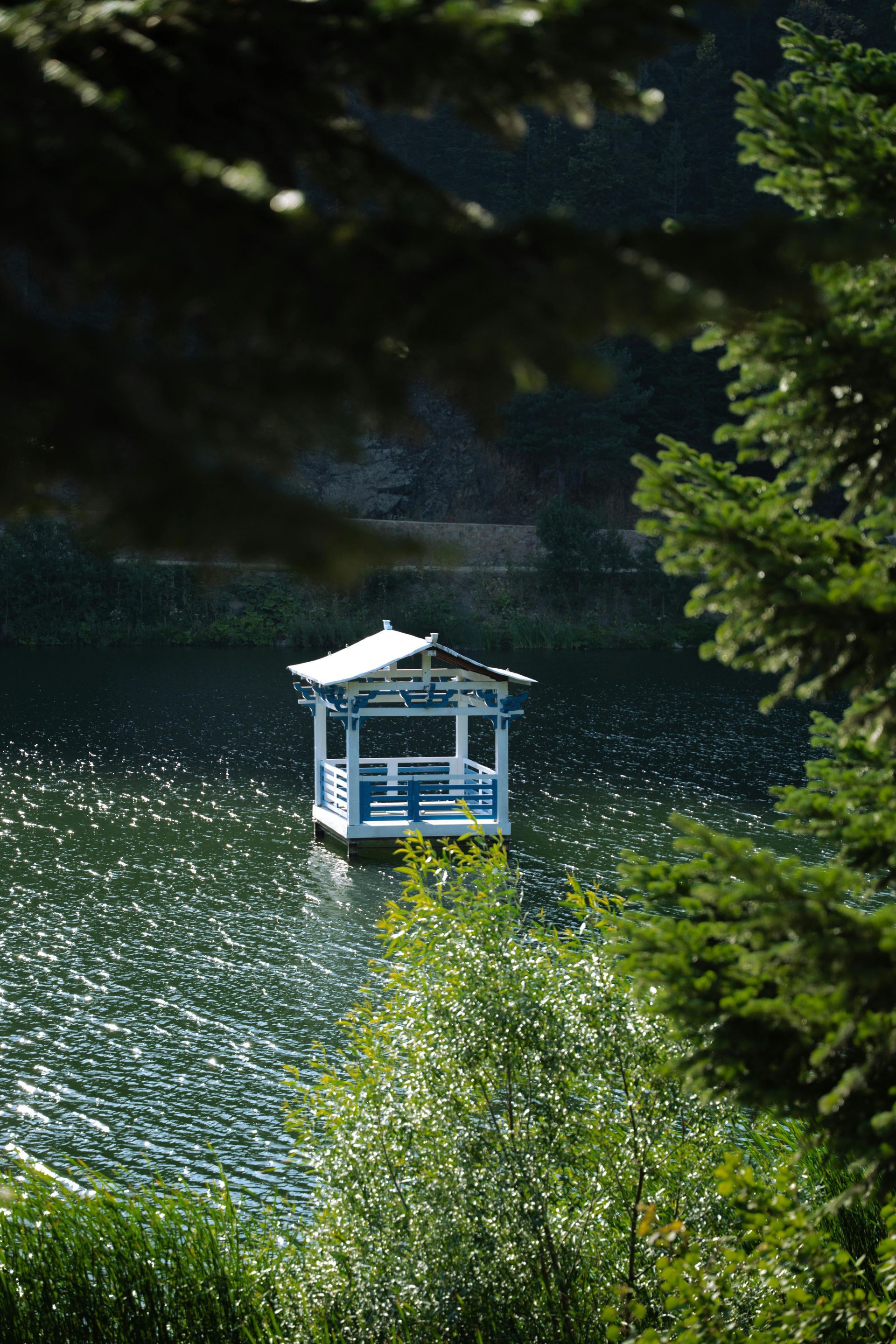 Blue and white gazebo on lake, framed by green trees.