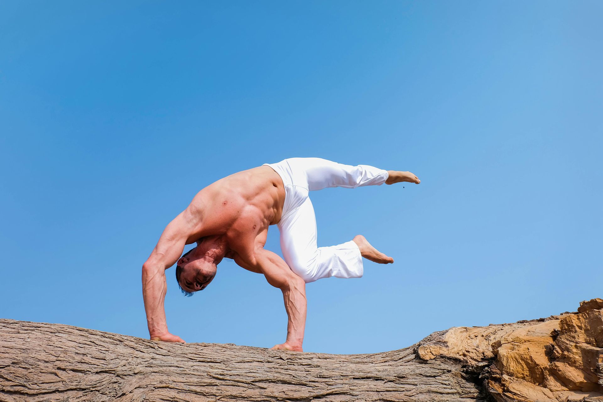 Man performing yoga pose on a rock, against a blue sky. He is shirtless and wearing white pants.
