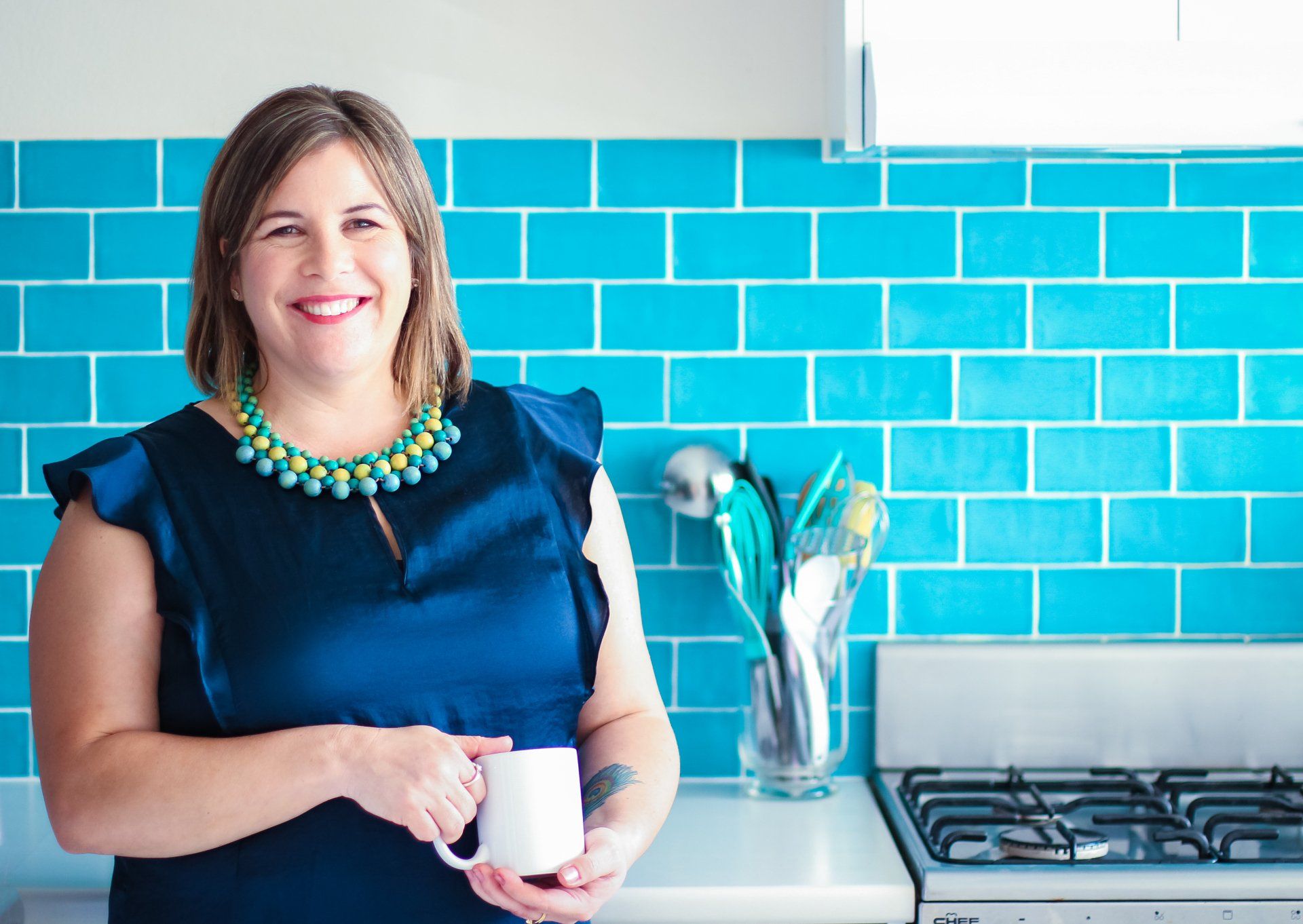 A woman with blue top in the kitchen with blue tiles in the background.