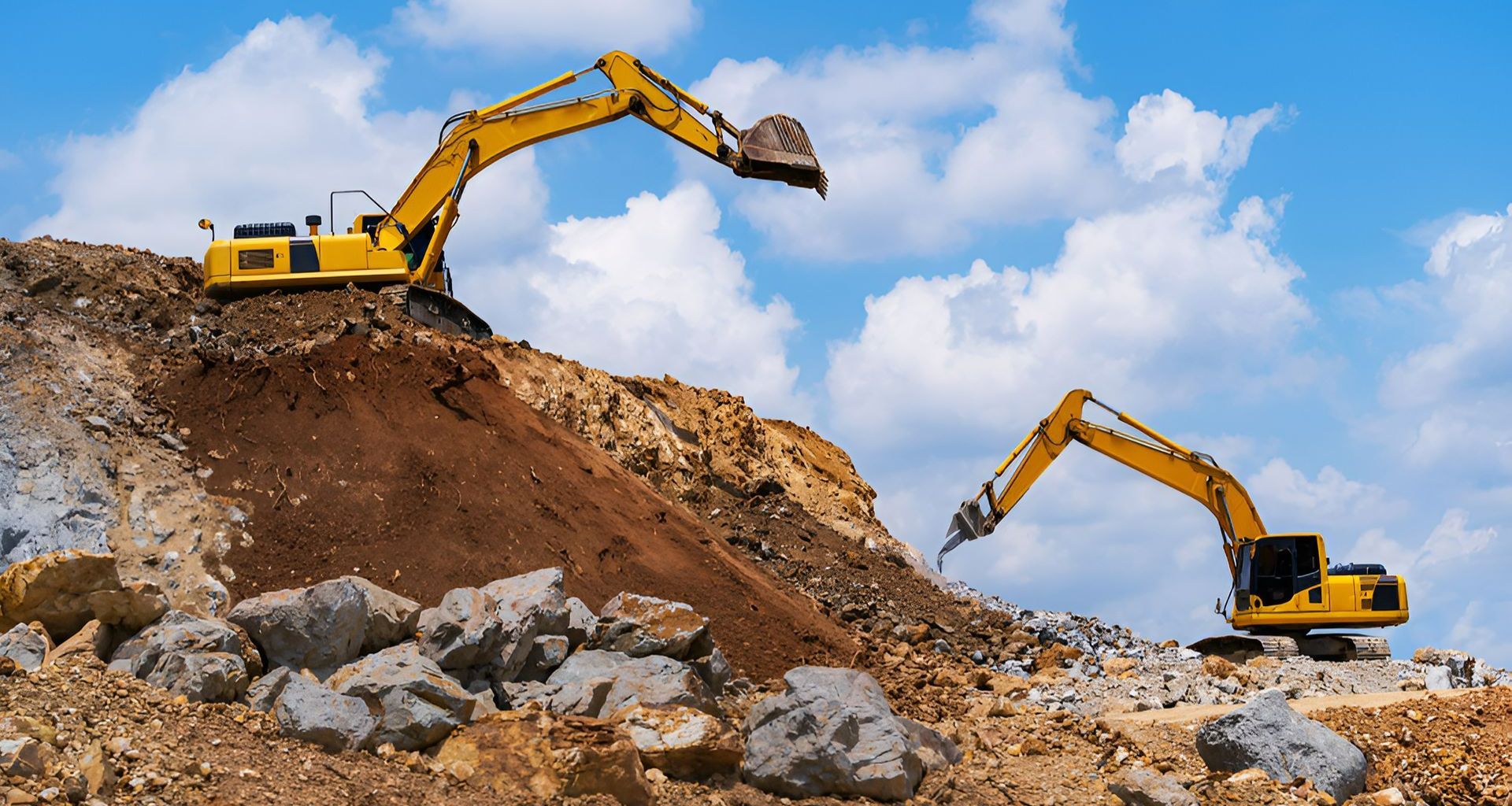 Two Yellow Excavators On A Rocky Hill Excavating Soil, Against A Blue Sky With Clouds