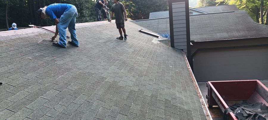 Two workers repairing a shingled roof beside a residential driveway and parked dumpster.